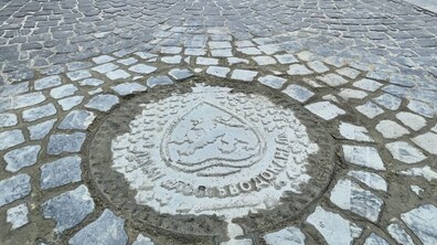 Крупним планом зображено фрагмент брукованої дороги. У центрі кадру — круглий люк, вмонтований у бруківку. Він має рельєфну поверхню та декоративний малюнок у вигляді герба. По краю люка є напис ЛМКП «Львівводоканал», виконаний по колу.