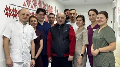 Усміхнені пацієнт Михайло та команда лікарів (дев’ятеро медичних працівників) стоять у холі лікарні святого Луки; стіна позаду розмальована узором вишиванки з червоно-чорним орнаментом