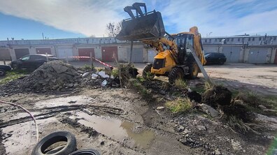 На фото зображена ділянка на відкритій території у центрі якої працює жовтий екскаватор-навантажувач. Його ківш піднятий угору і заповнений землею. Поряд видно ями, нахилений металевий стовп та знесений паркан з дроту