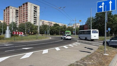 Міське перехрестя з кільцевим рухом, де на асфальті нанесено білу розмітку зі стрілками напрямку руху. У кадрі видно білий та зелений автобуси і білий легковий автомобіль, а на правому передньому плані - синій дорожній знак «Напрямки руху по смугах».