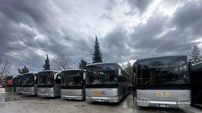 Вісім великих сріблястих автобусів стоять рівним рядом на відкритому майданчику на вологому від дощу асфальті під похмурим сірим небом. На кожному з них видно логотип італійської компанії TUA. На другому плані видніються дерева