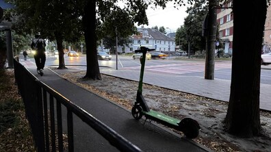 На вечірній вулиці на передньому плані стоїть прокатний електросамокат. Ліворуч доріжкою їде чоловік на електросамокаті. Позаду видно дерева, дорогу з автомобілями та будинки.