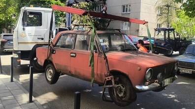 Евакуатор за допомогою спеціальних кріплень піднімає старий червоний автомобіль ВАЗ. На другому плані - багатоповерхівка та зелені дерева