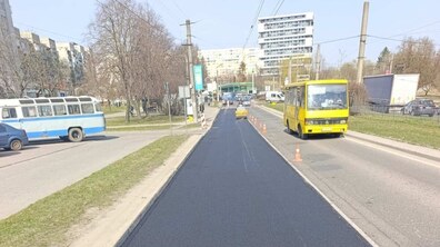 Міська вулиця під час дорожніх робіт у світлий день. У центрі зображення — свіжа смуга асфальту. Праворуч від неї рухається жовтий автобус, а вздовж смуги виставлені помаранчеві дорожні конуси, що відокремлюють зону ремонту від руху транспорту. На задньому плані — багатоповерхові житлові будинки та опори з контактними лініями для електротранспорту.