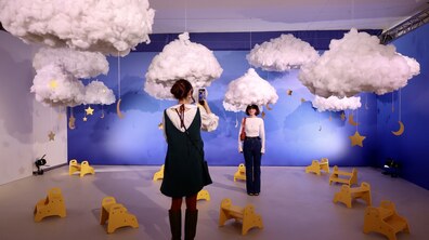 У виставковому просторі з синім тлом та підвішеними білими хмарами дівчина фотографує відвідувачку, яка позує під декоративним «небом». На світлій підлозі кімнати хаотично розставлені маленькі жовті стільчики.