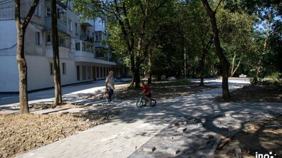 Нові алеї вимощені з бруківки у дворі будинку. Поміж алеями ростуть зелені дерева. Доріжки тестує хлопчик на двоколісному велосипеді. До нього на зустріч йде бабуся з сумкою в руках
