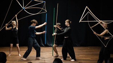 П’ятеро учасників постанови, одягнених у стриманий чорний одяг, босоніж, взаємодіють із дерев’яними конструкціями та довгими палицями, що нагадують геометричні форми, каркаси або абстрактні інструменти.