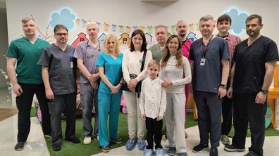 Спільне фото: пацієнт Юрчик з мамою Іриною та командою лікарів центру трансплантології та лікарні святого Миколая стоять у холі медичного закладу. Усі усміхнені й дивляться прямо в камеру. Позаду видно розмальовану стіну та декорації.
