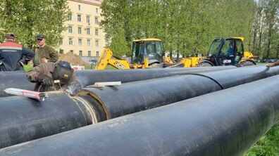 Тривають роботи із заміни тепломережі. На передньому плані лежать кілька великих чорних труб, розташованих паралельно одна до одної. Один із працівників у захисній масці зварює стик труби.