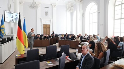 Офіційна зустріч із німецькою делегацією у Великій сесійній залі Львівської міської ради. У лівій частині кадру біля прапорів Німеччини, України та Львова стоїть Андрій Москаленко, перший заступник міського голови Львова, із мікрофоном у руках. Учасники заходу в діловому одязі сидять півколом за темними столами, слухають промовця та переглядають презентацію на великому екрані.
