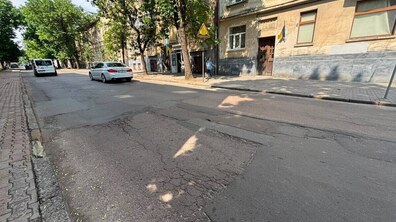 Ділянка вул. Героїв УПА. На передньому плані видно зношене асфальтне покриття з тріщинами, латками та вибоїнами. По дорозі їде кілька авто. Вздовж вулиці стоять кількаповерхові житлові будинки, ростуть дерева.
