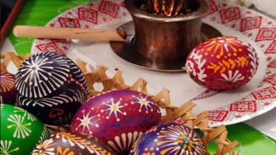 На передньому плані плетений кошик із яскравими великодніми писанками, розписаними традиційними візерунками (зірками, лініями та рослинними мотивами). Позаду на тарілці з орнаментом стоїть прилад для розтоплювання воску з писачком.