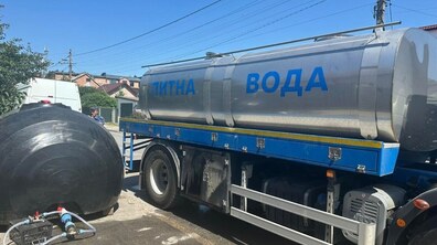 Праворуч стоїть велика автоцистерна з написом «Питна вода» синіми літерами. Поряд із нею розташована чорна пластикова ємність, до якої під’єднано шланги для закачування води.