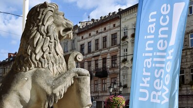 Кам’яна скульптура лева на площі Ринок у Львові, поруч із якою майорить блакитний прапор із написом «Ukrainian Resilience Week». На задньому плані видно історичні будівлі, вуличні ліхтарі та квіткові композиції