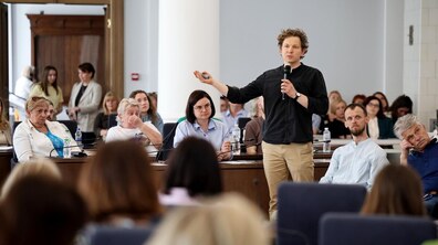 Антон Коломєйцев виступає перед слухачами. Зал наповнений людьми. Всі уважно спостерігають за спікером