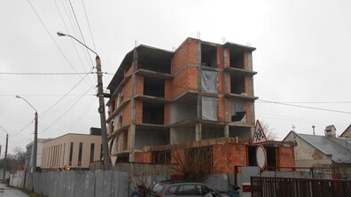П’ятиповерхова недобудована цегляна будівля огороджена металевим парканом. Всередині подекуди вікна закриті плівкою. Попереду припаркована автівка. Ходять люди.