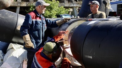 Роботи із заміни теплової мережі. На передньому плані працівник у захисній масці зварює великі чорні труби теплотраси. Поруч стоять двоє колег: один у робочому спецодязі та захисних рукавицях. Навколо видно великогабаритні труби, будівельні матеріали та техніку, а на задньому плані — конструкцію будівлі та зелені дерева.
