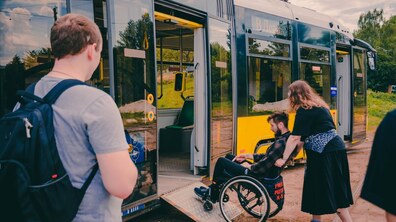 Фото: Володимир Караїм. Жінка допомагає чоловікові у кріслі колісному зайти по відкидному пандусу всередину сучасного низькопідлогового трамвая. На передньому плані спиною до кадру стоїть хлопець із рюкзаком та спостерігає за процесом.