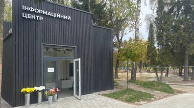 Сучасна споруда інформаційного центру темного кольору з відчиненими скляними дверима, біля яких стоять вази з квітами. На тлі за металевою огорожею видніється поле почесних поховань з українськими прапорами серед дерев.