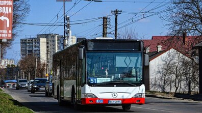 Великий низькопідлоговий автобус білого кольору з червоним бампером, який курсує за маршрутом №51 у Львові. Транспорт рухається міською вулицею, на другому плані - житлові будинки та автомобілі.