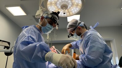 Двоє лікарів, одягненні у медичні халати, шапочки та маски, зі спеціальними операційними окулярами на голові, оперують в операційній, над ними – світить операційна лампа