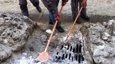 Зимовий тротуар, обабіч - скупчення підталого снігу та льоду. Комунальні працівники нахилилися і розчищають дощоприймач. Решітка темна, металева, з вертикальними отворами, частково вже очищена від снігу та льоду. З обох боків дощоприймача лежить сніг, що згребли під час очищення.