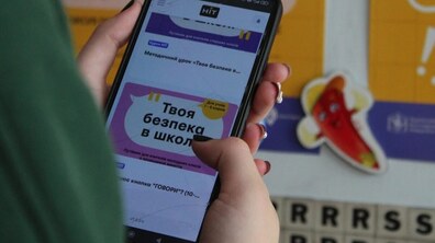 Учениця тримає в руках смартфон, на екрані якого відкритий навчальний матеріал «Твоя безпека в школі». На задньому плані видно яскраві навчальні плакати, що створюють атмосферу освітнього простору.