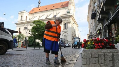 На передньому плані працівниця комунальної служби в помаранчевому жилеті підмітає мітлою бруковану міську вулицю. На другому плані видно історичну архітектуру — світлу будівлю храму, міські будинки та припарковану спецтехніку.