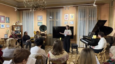 У затишній залі з класичним інтер’єром відбувається камерна подія — жінка виступає перед невеликою аудиторією, тримаючи в руках текст. Гості сидять на стільцях, уважно слухають. Поруч видно рояль і старовинний грамофон, що додають події особливого культурного настрою.