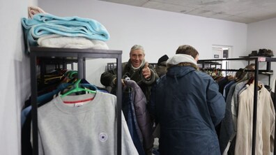 Усміхнений чоловік обирає собі верхній одяг серед стелажів з одягом. Поруч люди розглядають речі, які висять на вішалках, а на полицях зверху лежать складені ковдри. Фото ілюстративне.