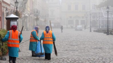 Три працівниці комунальних служб на площі Ринок, яка оповита туманом. Вони одягнені у спеціальні робочі жилетки, а в руках тримають робочий інвентар.