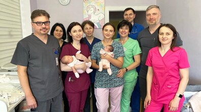 Команда медиків дитячої лікарні святого Миколая разом із двома маленькими пацієнтами після успішного лікування. Лікарі, медичні сестри та батьки стоять у лікарняній палаті, усі дивляться в камеру та усміхаються.