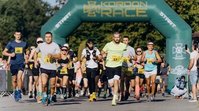 Група бігунів стартує під великою надувною аркою з написом «Kordon Race». Учасники у спортивному одязі з номерними знаками на грудях біжать по асфальтованій дорозі. Погода сонячна, навколо видно зелені дерева, а на задньому плані – інші учасники та глядачі (фото зі сайту kordonrace.com).