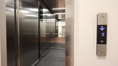 відкритий ліфт у сучасній будівлі. Кабіна ліфта має металеві блискучі стіни та поручень уздовж задньої стінки, підлога темна. Праворуч на стіні біля входу видно панель виклику ліфта з підсвіченим дисплеєм: світиться стрілка вниз і цифра «2», що означає, що ліфт їде вниз з другого поверху. Усередині кабіни та в коридорі — яскраве рівне освітлення.