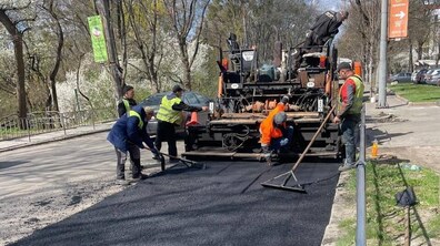 Ремонт дорожнього покриття. На передньому плані — свіжа смуга темного асфальту, яку щойно укладають. Позаду стоїть велика дорожня машина-асфальтоукладач, з якої рівномірно подається гарячий асфальт. Поруч працює кілька дорожніх робітників у яскравих жилетах — жовтих і помаранчевих.