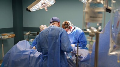Троє лікарів працюють за операційним столом - всі одягнені у медичні халати, шапки і маски; над лікарями світять дві операційні лампи