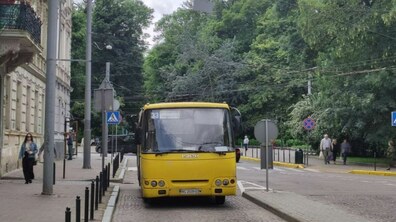 Жовтий автобус №33 стоїть біля тротуару на вузькій вулиці, ліворуч видно історичну будівлю, праворуч — густі зелені дерева, а неподалік ідуть пішоходи.