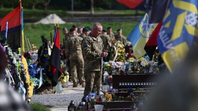 На Личаківському кладовищі військовослужбовець стоїть біля могили полеглого воїна: він схилив голову, замислено дивиться на могилу, в руках тримає берет. Позаду видно декількох військових, поруч – інші могили воїнів у прапорах.