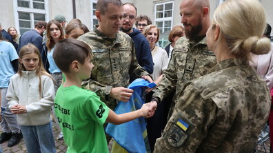 Хлопчик тисне руку усміхненому військовому. Поруч стоїть інший військовий з прапором України. Навколо - діти.
