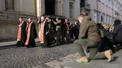 Похоронна процесія біля Гарнізонного храму: кілька священнослужителів йдуть попереду, один із них тримає хрест. За ними військові у камуфляжній формі несуть труну. Поруч ідуть люди в темному одязі, також люди поруч на колінах віддають шану.