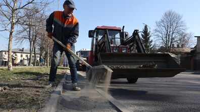 Працівник комунальної служби у робочому одязі та рукавицях тримає лопату із набраним піском, а поруч стоїть невеликий трактор із ковшем для завантаження сміття. Уздовж дороги ростуть дерева без листя, а вдалині видно житлові будинки та перехожих.
