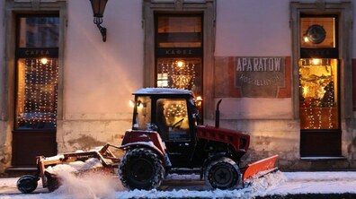 Невеликий червоний трактор прибирає сніг на міській вулиці. Спереду в нього встановлена широка лопата, якою він відсуває мокрий сніг убік. На тлі — будівля з освітленими вікнами кафе
