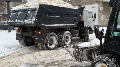 Зимова міська вулиця під час прибирання снігу. У центрі фото — велика вантажівка з відкритим кузовом, повністю заповненим снігом. Сніг насипаний високою купою, вище бортів. Праворуч видно іншу снігоприбиральну машину з широким металевим відвалом, який згрібає брудний сніг із проїжджої частини. На задньому плані — багатоповерхові житлові будинки з вікнами та балконами, фасади сірі, типові для міської забудови.
