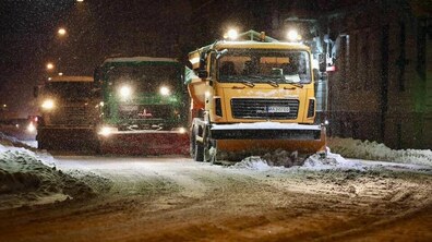 Нічна зимова вулиця Львова; по засніженій дорозі їде три піскороскидачі, один за одним, з увімкненими яскравими фарами. Кожна машина має спереду відвал для розчистки снігу. Дорога вкрита снігом, з боків – високі снігові кучугури.