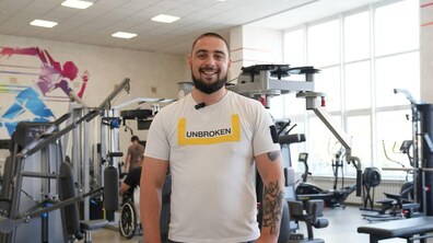 Захисник Володимир Пономарчук усміхається, стоячи у залі фізичної реабілітації центру UNBROKEN. Він одягнений у футболку з логотипом UNBROKEN, а на задньому плані видно тренажери та іншого пацієнта.