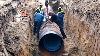 Крупним планом показано, як працівники Львівводоканалу в глибокій траншеї, виритій у ґрунті, монтують нову водопровідну трубу. Один робітник у зварювальній масці зварює стик на великій металевій трубі, що лежить у траншеї. Ще троє працівників у захисних жилетах стоять поруч. Всередині металевої труби є синя пластикова труба.