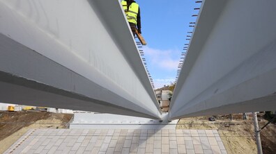 Монтаж мостових балок. У центрі кадру під кутом сходяться дві масивні сірі мостові балки, між якими у вузькій щілині видно робітника в яскраво-жовтому світловідбивальному жилеті та кепці. На задньому плані під балкою – вимощений схил та будівельний майданчик.
