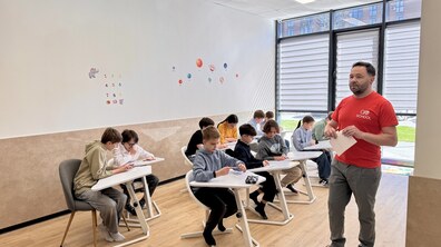 Близько десятка учнів сидять за партами у світлому класі та дивляться на завдання на листках. Біля них вчитель у червоній футболці з емблемою школи. На білій стіні позаду зображені цифри та планети сонячної системи