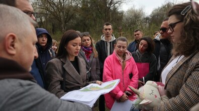 Кілька десятків мешканців стоять в колі під час зустрічі щодо будівництва житла. Посередині жінка тримає в руках карту з проєктом житлового комплексу та розповідає деталі