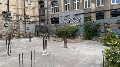 Занедбана ділянка між старими будівлями. На бетонному майданчику видно металеву арматуру від незавершеного будівництва, довкола — металевий паркан із графіті та зарості кущів.